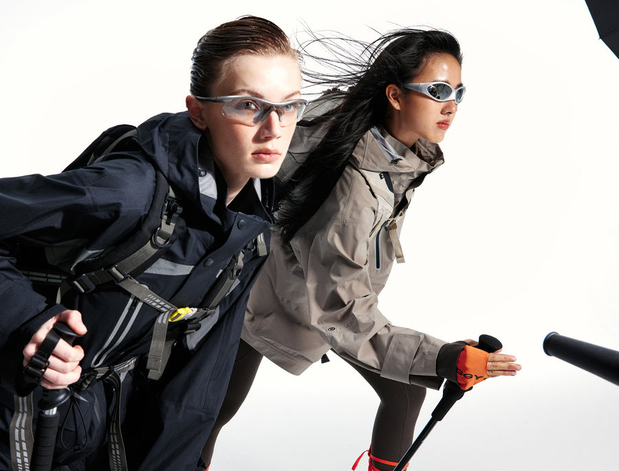 Two women in FENGGY technical jackets lean into forward motion, side by side, eyes shielded by sport shades against a soft studio light.