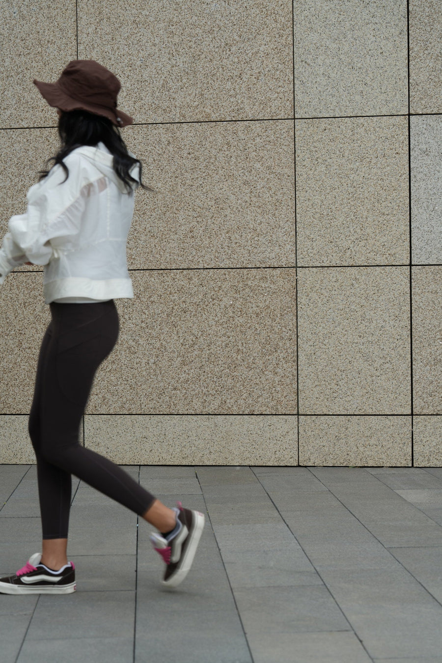 A figure in a white TITTALLON shell jacket mid-stride past a pale stone wall in daylight.