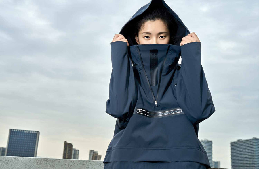 A figure in a navy TITTALLON Urban Survival technical shell, hood drawn up and collar high, against an overcast city skyline.