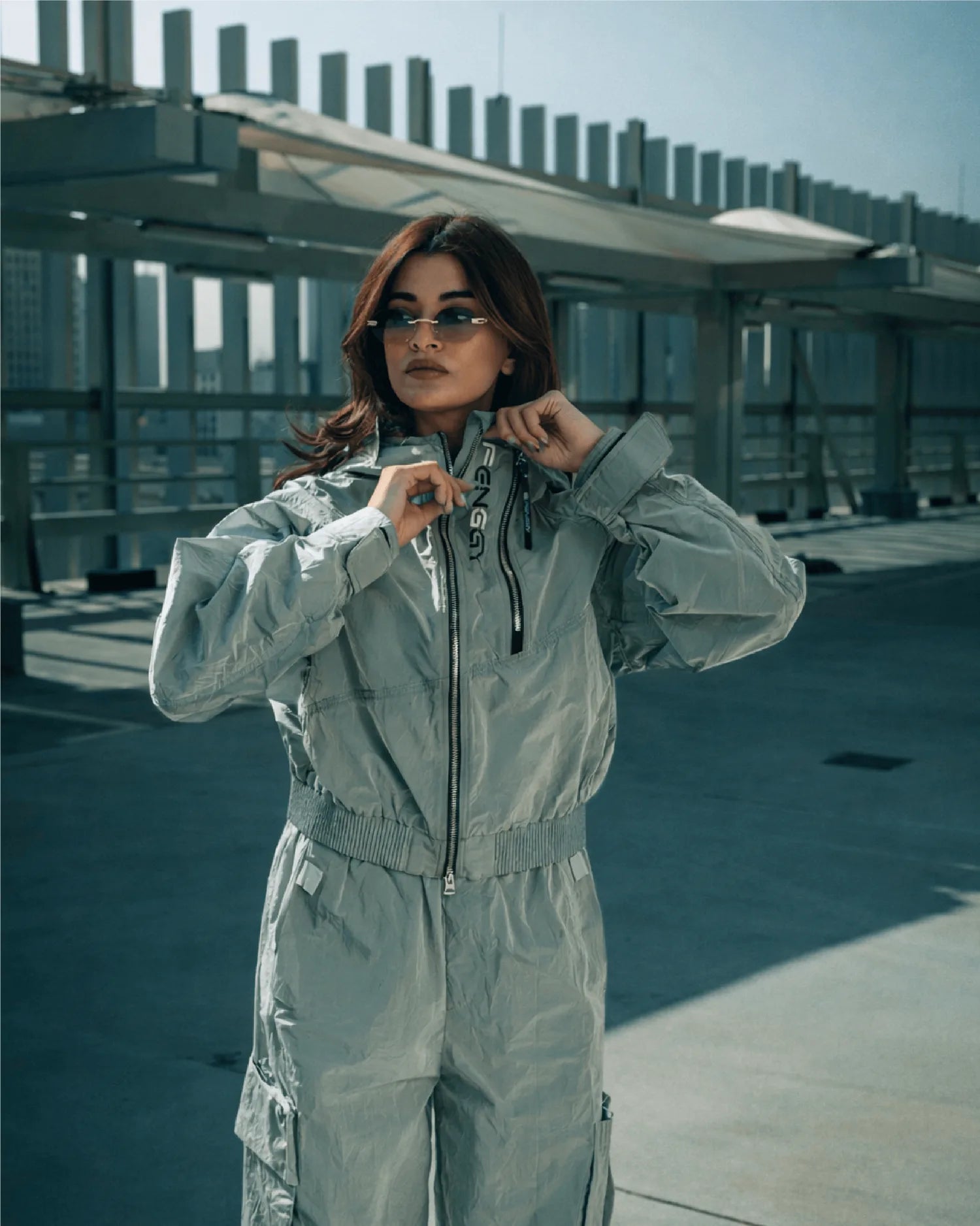 A composed woman in a grey FENGGY crinkle-nylon jumpsuit on an open rooftop at edge-of-morning, slatted metal railings catching cool light behind her.