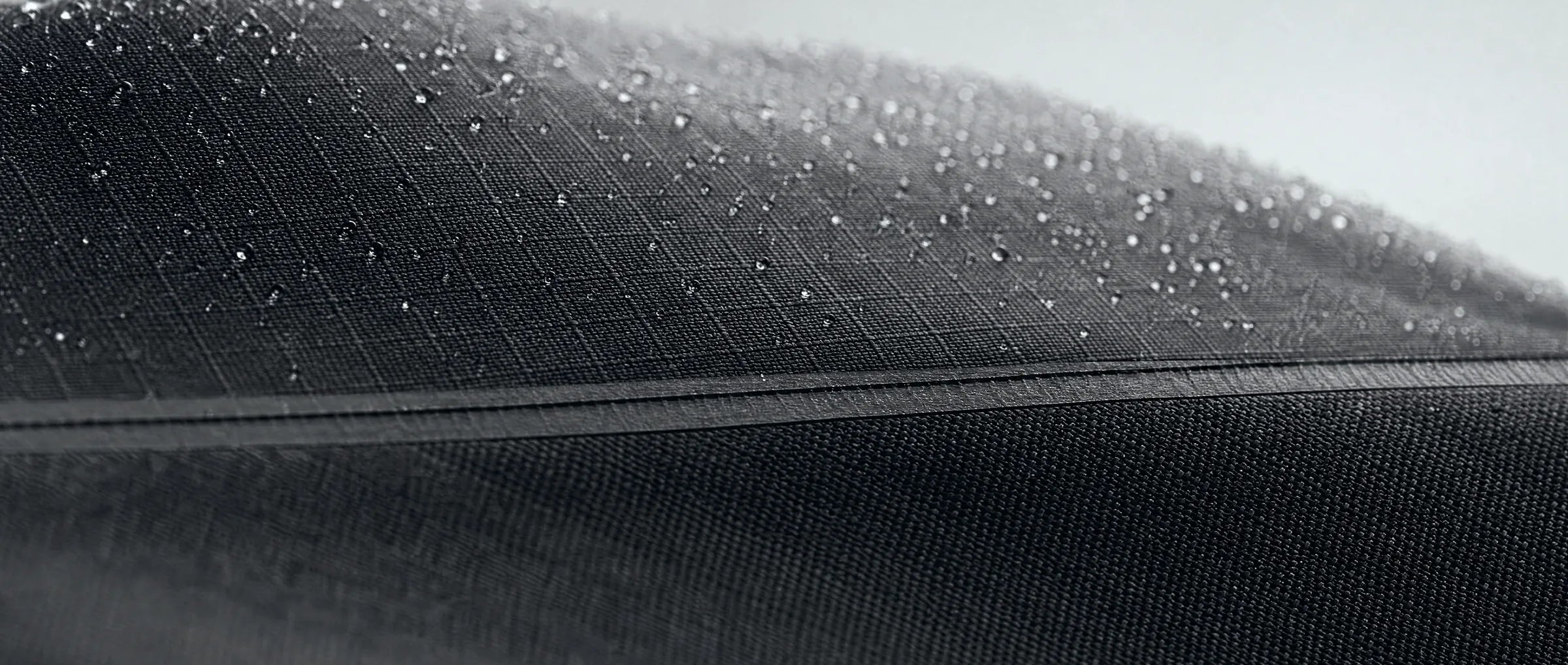 Extreme close-up of a graphite technical shell fabric folded into a low ridge, a bonded seam crossing it, water beading on the weave