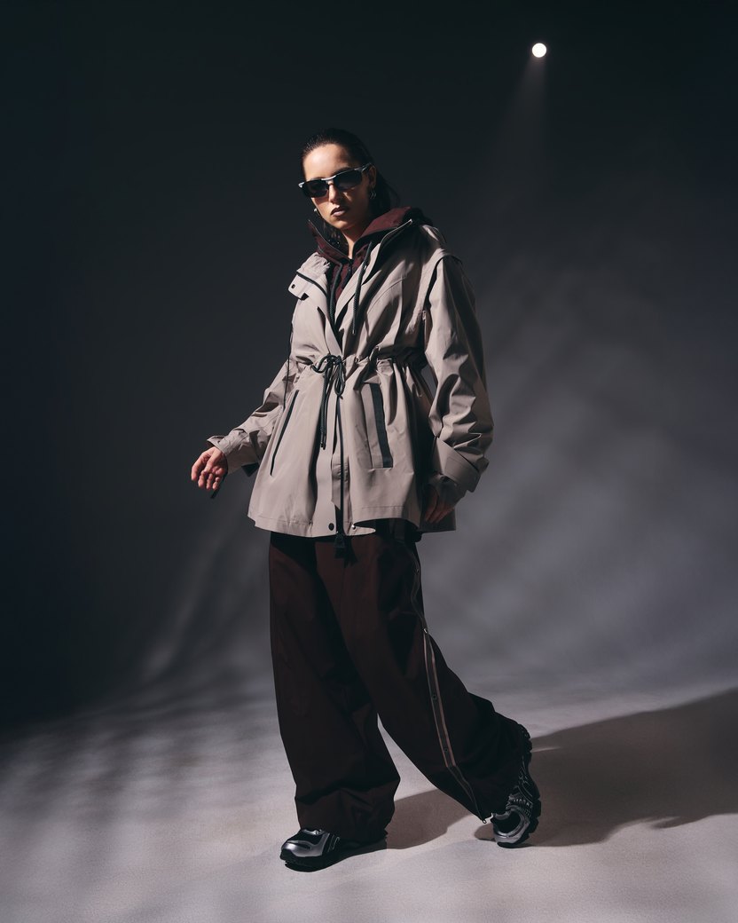 A woman in a taupe hooded parka and wide pants under a spotlight